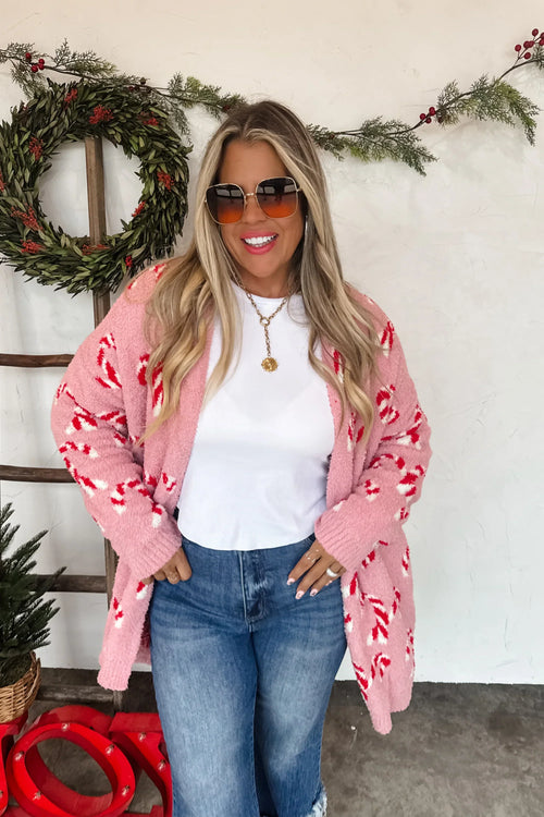 Woman wearing a pink cardigan with candy cane pattern, white shirt, and blue jeans in front of a Christmas wreath.