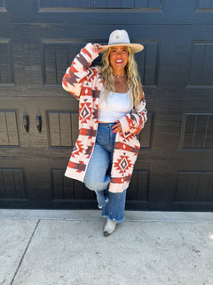 A woman stands in front of a black garage door wearing a cream and rust Southwestern patterned cardigan, a white tank top, blue jeans, a beige wide-brim hat, and gray boots.