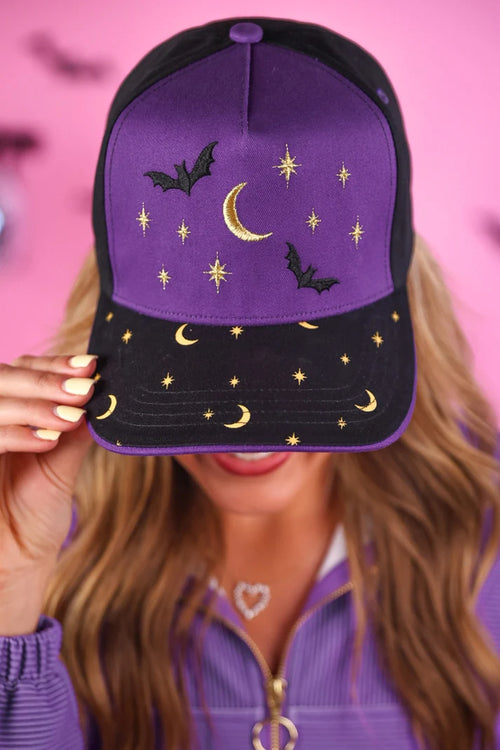 Purple cap with star, moon, and bat designs held by a person against a pink background