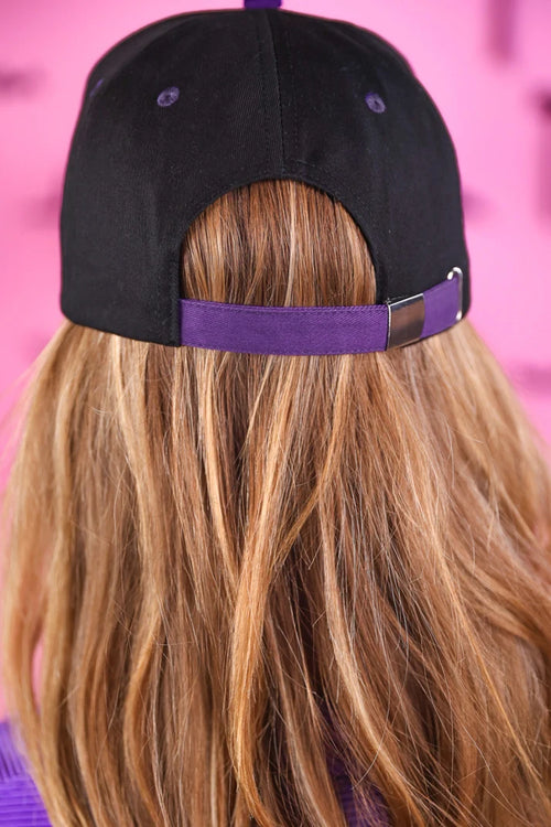 Back view of a person with long blonde hair wearing a black baseball cap with a purple strap and matching purple eyelets, set against a pink background.