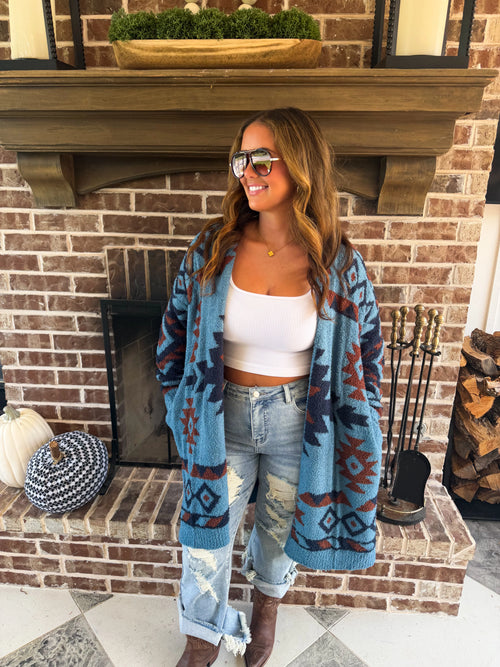 A woman stands in front of a brick fireplace wearing a blue, brown, and red geometric pattern knit cardigan over a white crop top and light-wash jeans.