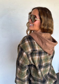 A woman wearing a brown and green plaid hooded flannel shirt over a tan hoodie, styled with gold-framed sunglasses, posing against a textured white wall.