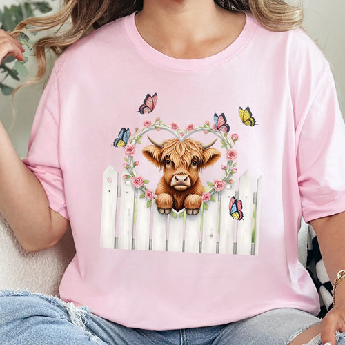 Soft pink graphic tee featuring a highland cow peeking over a white fence with pink florals and butterflies.
