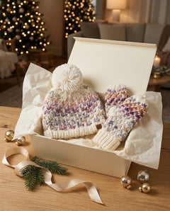 A cream knit beanie with a matching pom-pom and coordinating knit mittens, displayed inside an open cream gift box on tissue paper. The scene is styled on a wooden table with holiday greenery, gold ornaments, and a softly blurred living room background with lit Christmas trees.