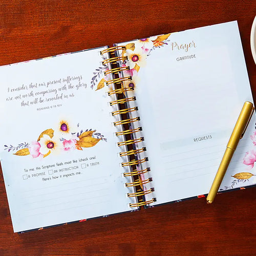 Open planner with floral design on a wooden surface, featuring a gold pen.