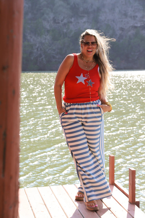 Woman in a red top with star designs and blue and white striped pants standing on a wooden dock by a body of water.