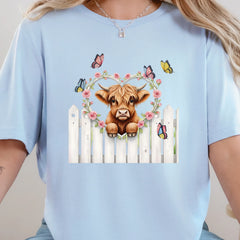 Baby blue graphic tee featuring a highland cow peeking over a white fence with florals and butterflies.