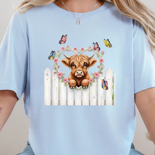 Baby blue graphic tee featuring a highland cow peeking over a white fence with florals and butterflies.