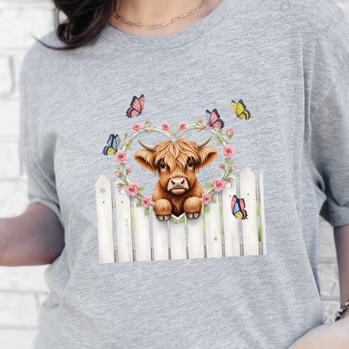 Gray graphic tee featuring a highland cow peeking over a white fence with florals and butterflies.