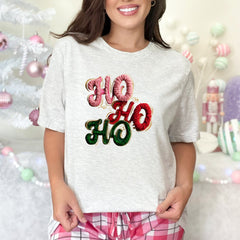 Person wearing a white t-shirt with 'HO HO HO' text in festive colors, standing in front of a decorated Christmas tree.