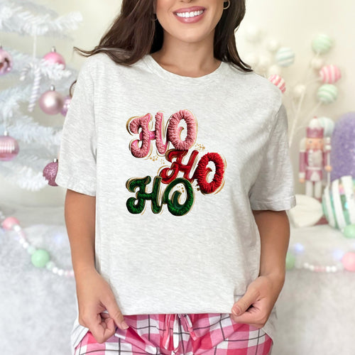 Person wearing a white t-shirt with 'HO HO HO' text in festive colors, standing in front of a decorated Christmas tree.