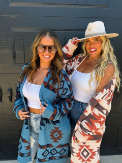 Two women posing in front of a dark gray door, showcasing two different Southwestern-style knit cardigans. The woman on the left wears a blue cardigan with brown and red geometric patterns over a white crop top and jeans.