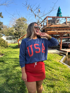 Model wearing a red fleece-lined jogger skirt with a white drawstring, paired with a blue USA sweatshirt on a green lawn.