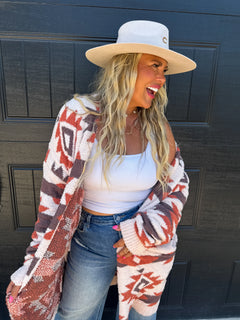 A woman wearing a white wide-brim hat, a white tank top, blue jeans, and a chunky knit cardigan with a red, brown, and cream Southwestern geometric pattern, standing in front of a black paneled door.