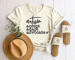 Beige t-shirt with 'Autism Accept. Adapt. Advocate.' text, tan hat, and sandals on a light wooden surface.