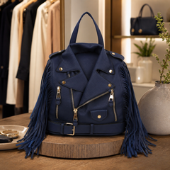 A navy blue vegan-leather biker jacket backpack with long fringe details and gold-tone hardware, displayed on a wooden pedestal in a boutique setting.