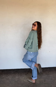 Woman wearing a green jacket and blue jeans standing against a white wall.
