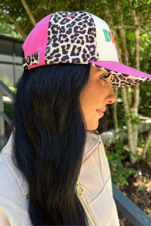 A woman in profile wearing a colorful trucker-style baseball cap with leopard print, pink panels, and a white front panel with a green letter, photographed outdoors.