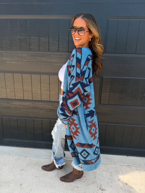 A woman wearing a blue knit cardigan with a red and dark gray geometric pattern, standing in front of a dark garage door.