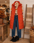 A styled autumn outfit displayed on a hanger against a white shiplap wall, featuring a bright orange quilted puffer vest over a long-sleeve floral sweater with pumpkin motifs, blue jeans, black ankle boots, and a woven scarf on a wooden ladder.