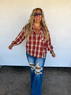 A woman modeling a red and beige plaid button-up shirt with blue ripped jeans, standing against a white stucco wall.