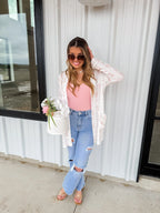 Full length view of a woman wearing a white waffle-knit 'Lola' cardigan featuring a pink bunny print, styled with a pink tank top and distressed denim jeans.