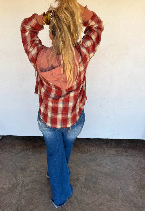 A woman viewed from behind wearing a red and white plaid flannel shirt with a peach-colored hood, blue jeans, and gold bracelets, standing against a white wall.