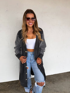 Woman wearing a long dark gray cardigan, white top, and ripped jeans against a white wall.