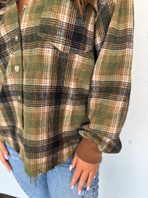 A close-up of a person wearing a long-sleeve plaid button-up shirt in olive green, tan, and black, paired with light blue jeans.