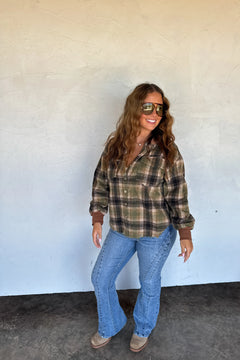 A woman wearing a green and brown plaid flannel shirt, blue jeans, and brown boots stands against a textured off-white wall.