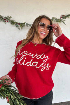 Woman wearing a red sweater with 'Howdy Holidays' text, standing against a white wall with greenery.