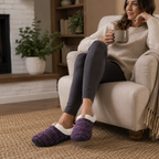 A woman sits in a beige armchair in a cozy living room, wearing a knit purple slipper with a white fuzzy trim and a small gold bow. She is also wearing dark gray leggings and a cream ribbed sweater, holding a gray mug.