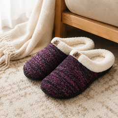A pair of knit slip-on house slippers with a cream sherpa lining, placed on a beige carpet next to a wooden chair and a cream throw blanket.