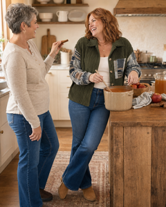 Two women laughing in a warm, rustic kitchen. The woman on the right wears a green ribbed vest over a plaid long-sleeve shirt, a white top, and blue jeans, holding a wooden spoon in a bowl.