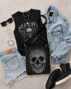 Black t-shirt with skull design, denim jacket, shorts, black sneakers, and studded skull bag on a light gray surface.