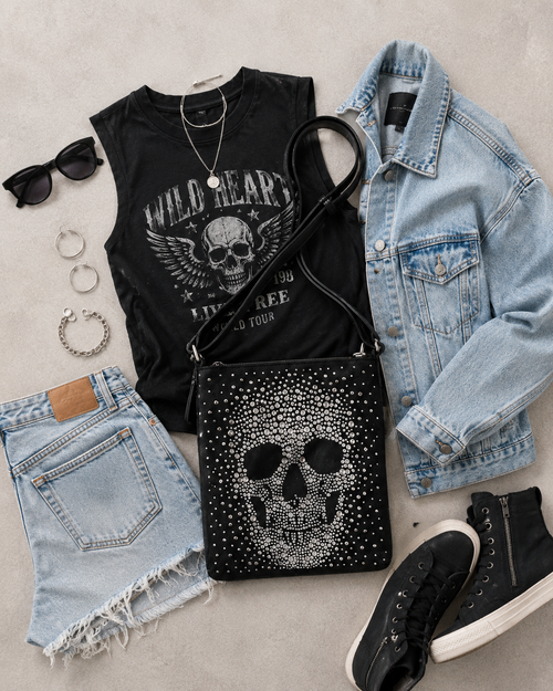 Black t-shirt with skull design, denim jacket, shorts, black sneakers, and studded skull bag on a light gray surface.