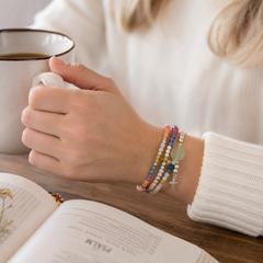 Woman wearing the Messy Life Charm Bracelet while holding a coffee mug beside an open devotional book in a cozy quiet-time setting.