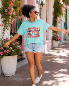 Woman wearing a light blue t-shirt with 'Salty Beach' design, standing outdoors with floral decorations.