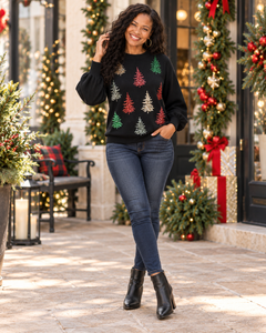 A woman stands outdoors in front of a decorated storefront wearing a black long-sleeve crewneck sweater with a festive pattern of red, green, and gold Christmas trees, paired with dark blue skinny jeans and black ankle boots.