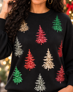 A close-up of a person wearing a black crewneck sweater decorated with sequined Christmas tree shapes in red, green, and silver, set against a softly blurred holiday background.