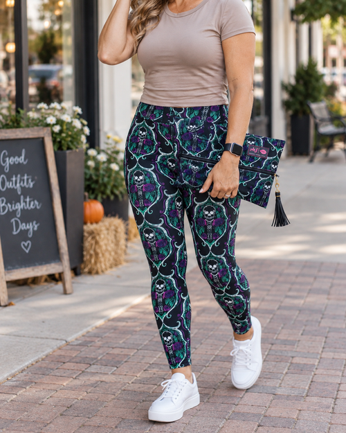 A woman wearing high-waisted leggings with a dark teal, purple, and black gothic skull and vine pattern, styled with a taupe t-shirt, white sneakers, and a matching tassel clutch on a brick sidewalk.