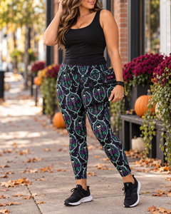 A woman wearing black and green patterned leggings with a skull-and-snakes design, styled with a black tank top and black sneakers, standing on an outdoor sidewalk with autumn leaves and pumpkins.