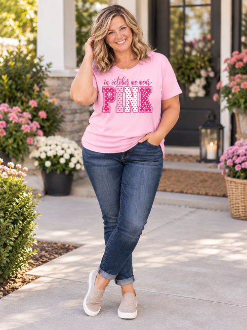A woman stands on a front porch wearing a light pink graphic T-shirt with the text 'in october we wear PINK' in pink and white, paired with dark blue jeans and beige slip-on shoes.