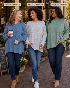 Three women walking outdoors on a brick sidewalk in front of a boutique, each wearing a different color of the same oversized long-sleeve sweater. Text labels at the top identify the models and sizes: Rita (5'6") wearing L/XL in blue, Maya (5'9") wearing S/M in light gray, and Simone (5'9") wearing 2XL in sage green.