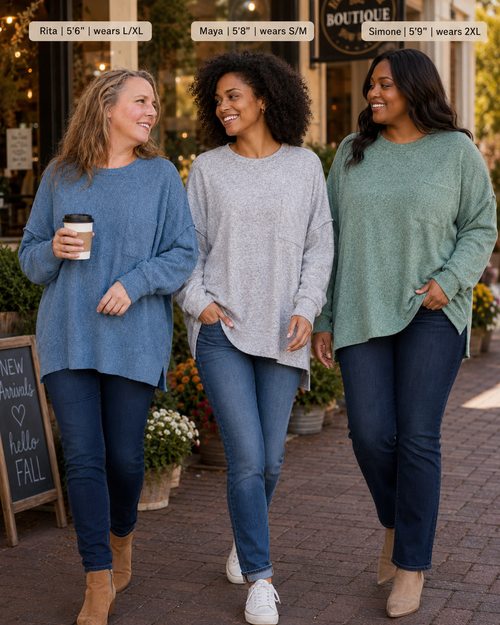Three women walking outdoors on a brick sidewalk in front of a boutique, each wearing a different color of the same oversized long-sleeve sweater. Text labels at the top identify the models and sizes: Rita (5'6") wearing L/XL in blue, Maya (5'9") wearing S/M in light gray, and Simone (5'9") wearing 2XL in sage green.