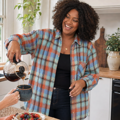 A woman in a kitchen wearing a blue, orange, and beige plaid button-up shirt over a black top, smiling as she pours coffee from a glass carafe into a dark mug.