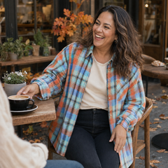 A woman smiling at an outdoor cafe table, wearing a long-sleeve plaid button-up shirt in orange, blue, and white over a cream t-shirt and black jeans.