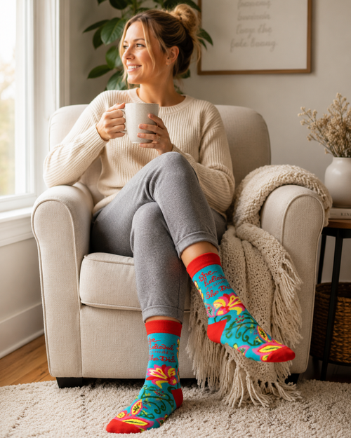 Woman sitting in a cozy armchair by a window holding a mug and wearing bright turquoise floral crew socks with red accents and the phrase She Believed She Could, So She Did.