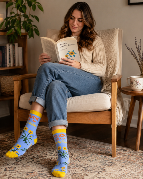 Woman sitting in a cozy reading chair with cuffed jeans, wearing blue and yellow floral Suck It Up Buttercup novelty crew socks.
