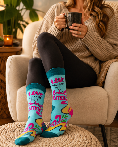 Woman relaxing in a cozy chair wearing teal Love Yourself You Badass Bitch crew socks while holding a black mug.